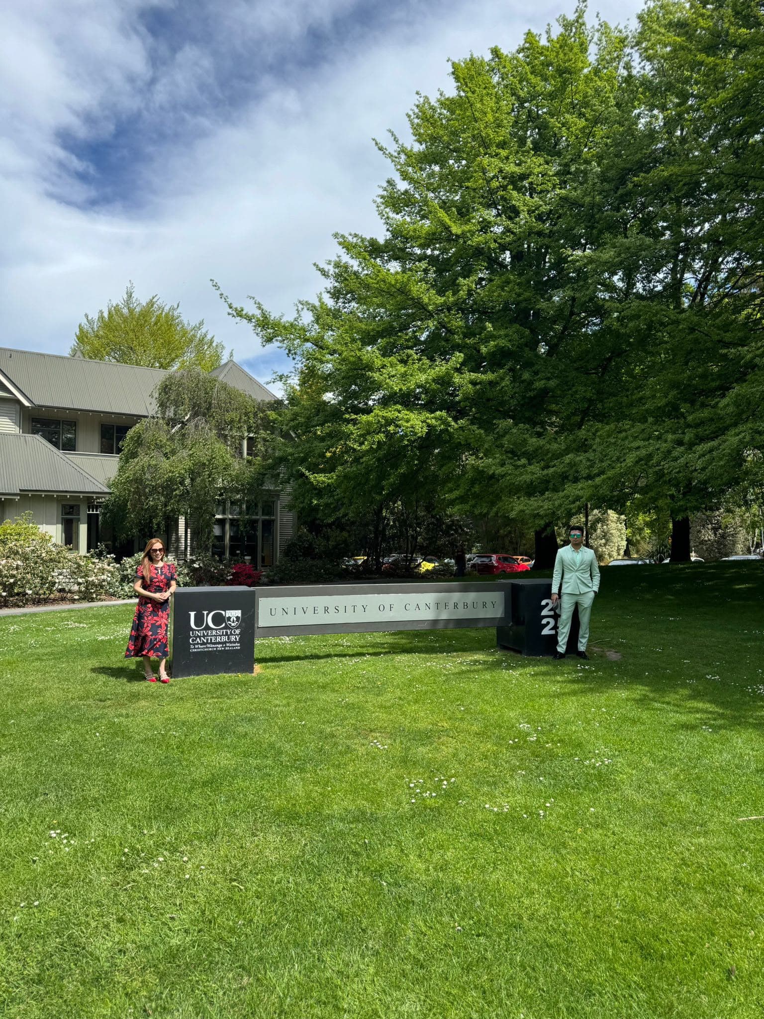 Two visitors beside the University of Canterbury sign on a green campus lawn